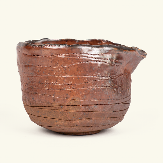 A handmade ceramic tea bowl with a rustic, textured surface and a small pouring spout, shown against a plain off-white background.