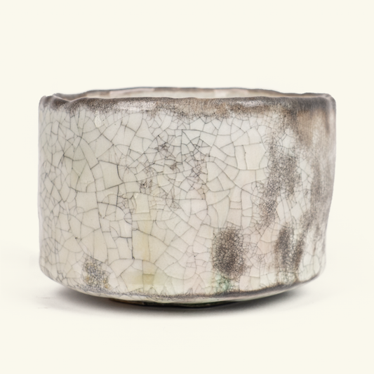 A handmade ceramic bowl with a wide, flared rim, featuring a glossy off-white glaze with a prominent crackle pattern and dark brown speckled accents, set against a plain cream background.
