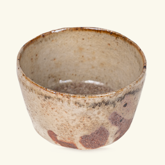 A handmade ceramic tea bowl with a rustic, hand-painted glaze in earthy beige, brown, and cream tones, shown against a solid off-white background.