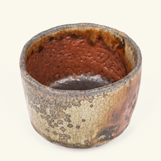 A handmade ceramic cup with a textured, speckled glaze in earthy brown, beige, and black tones, photographed against a plain beige background.