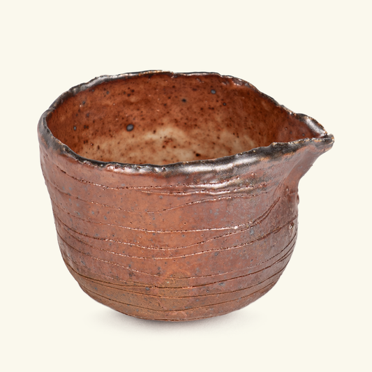 A handmade ceramic bowl with a rustic, hand-rolled rim and horizontal ridges on the body, featuring a warm reddish-brown glaze with dark brown speckles, set against a solid off-white background.