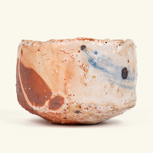 A handmade ceramic bowl with a rustic, organic shape, featuring a speckled beige and white glaze with a prominent brown patch on one side and blue swirls on the other, set against a solid off-white background.